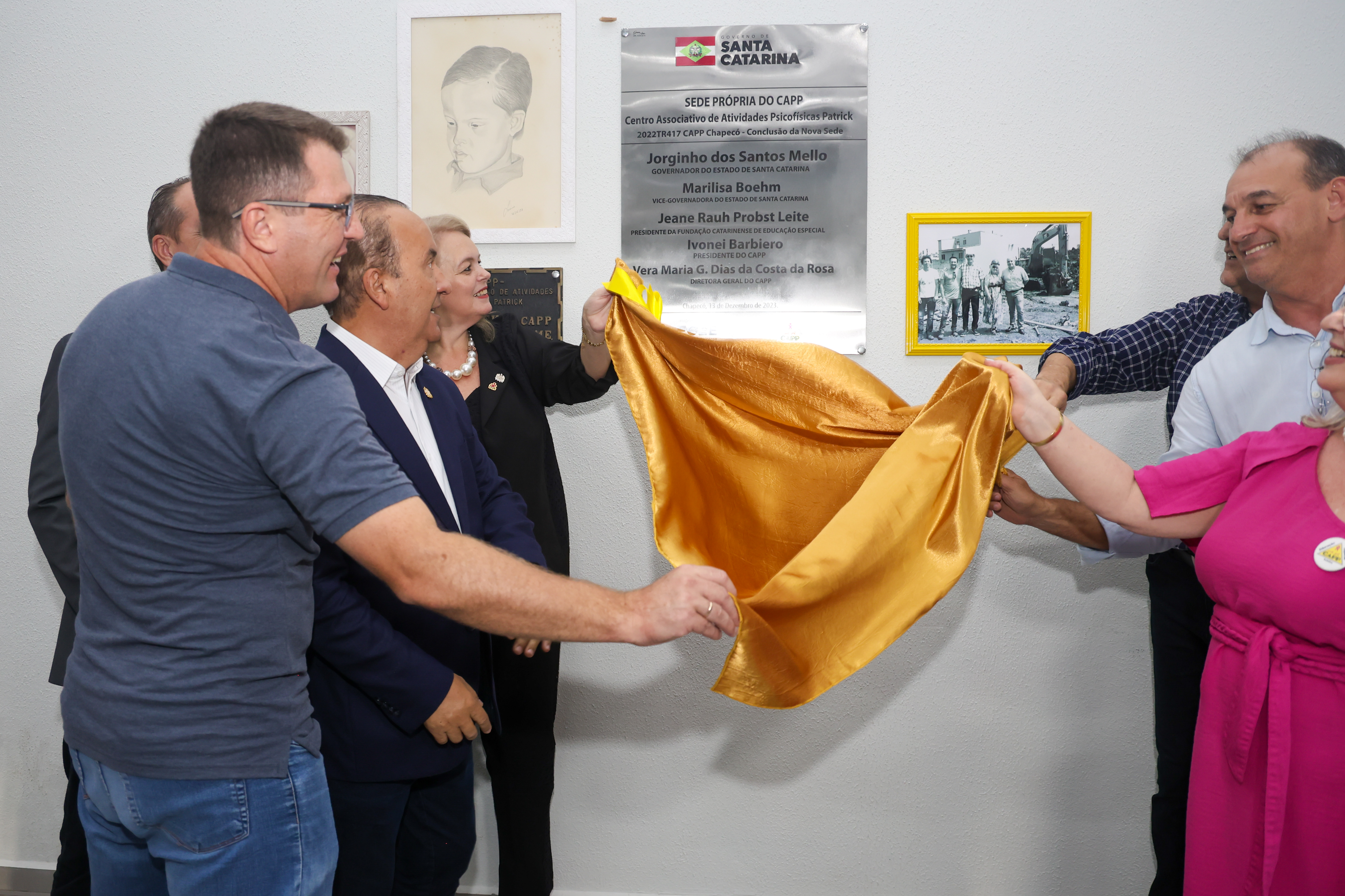 A imagem mostra sete pessoas discerrando a placa de inaguração do Centro Associativo de Atividades Psicofísicas Patrick (CAPP), em Chapecó. Entre as pessoas, o governador Jorginho Mello. Um pano amarelo em seda desce da placa de inauguração. Ele está sendo segurado pela autoridades que estão na imagem. No grupo apenas duas mulheres brancas, que estão nas extremidades da imagem. Ao fundo tem uma placa de inaguração onde se lê: Sede Própria do CAPP - Centro Associativo de Atividades Psicofísicas Patrick (CAPP).