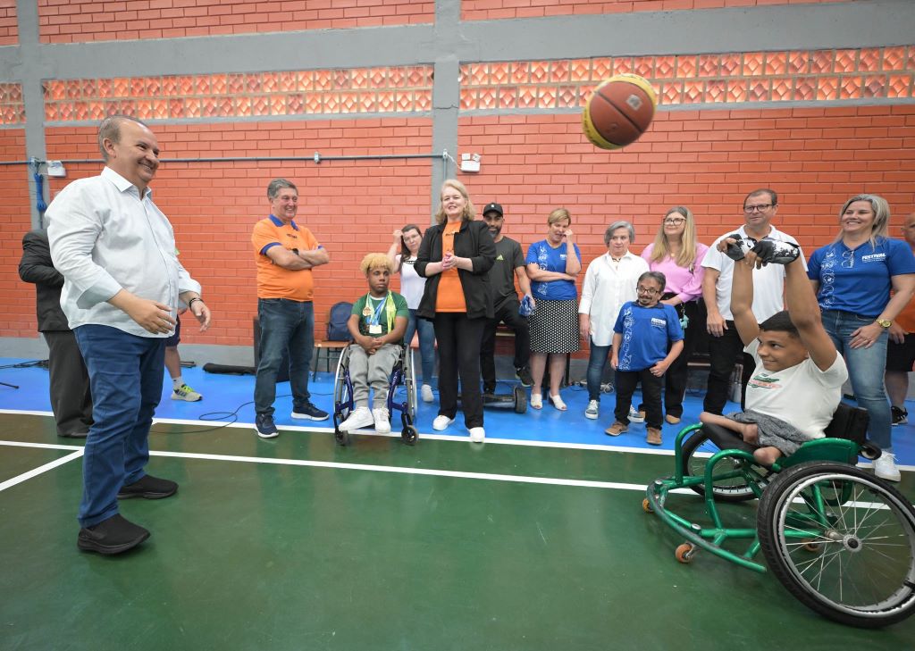 Foto: Eduardo Valente/ GOVSC Ginásio de esportes, homem em pé (Governador) joga bola com cadeirante, ao fundo pessoas assistem