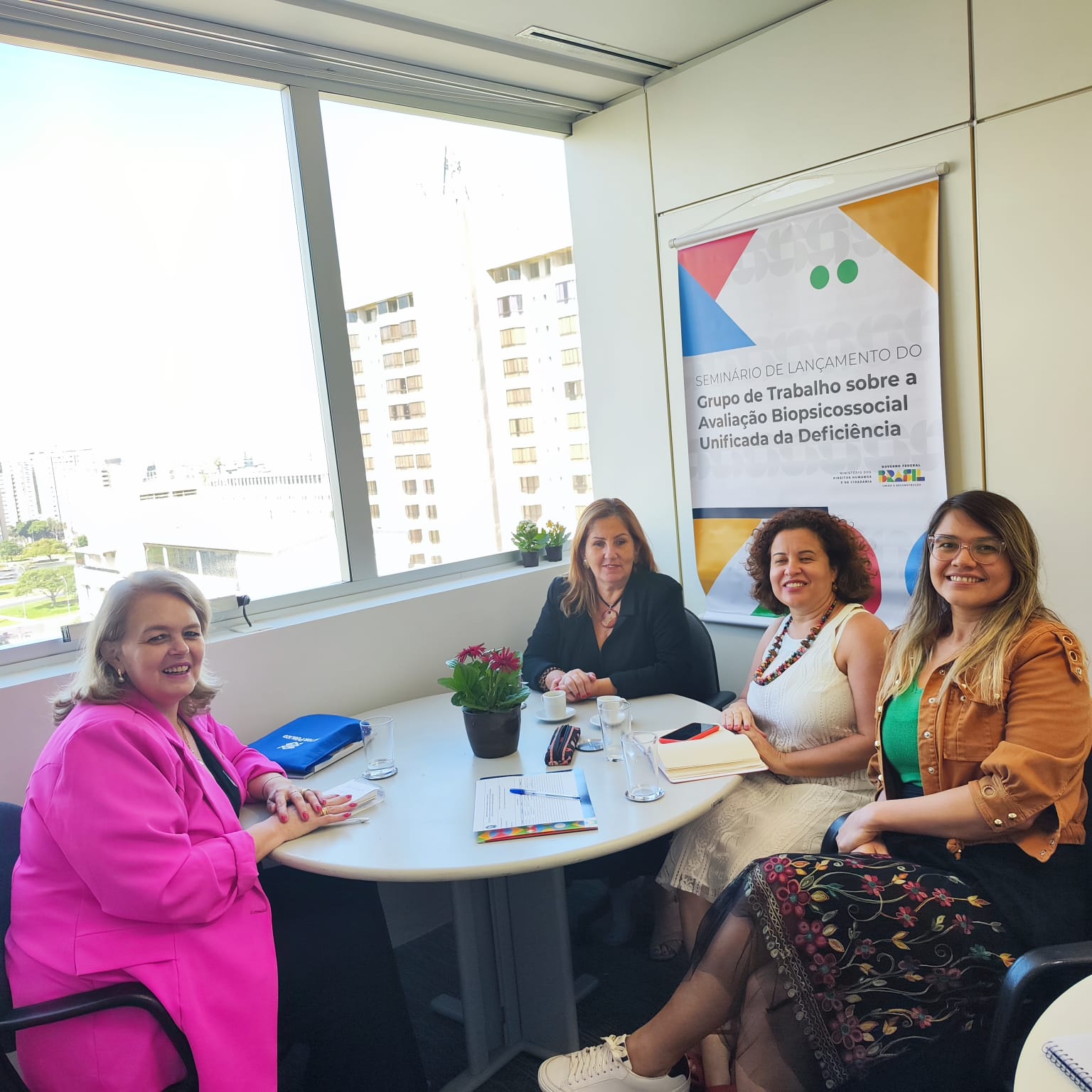Presidente da FCEE em Brasília com equipe da Secretaria Nacional dos Direitos da Pessoa com Deficiência 4 mulheres sentadas, sala de reunião, ao fundo banner com texto - Grupo de Trabalho sobre a Avaliação Biopsicossocial Unificada da Deficiência