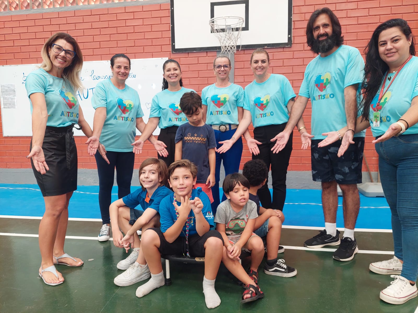 Evento do CETEA no ginásio da FCEE 15 pessoas em pé posam para foto em ginásio, 7 em pé vestidas com camisetas azuis com o símbolo do autismo, e abaixadas 8 crianças.