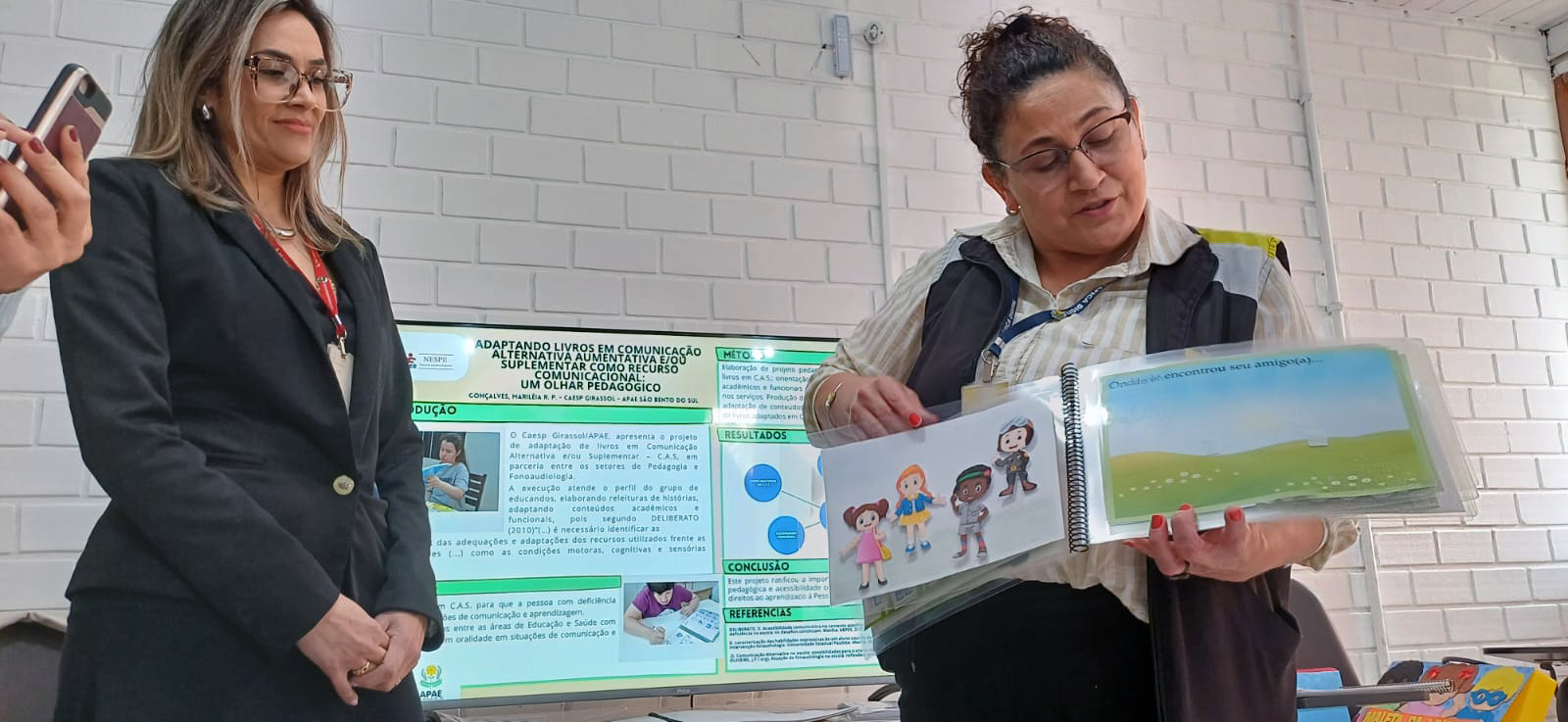 Professora da Apae de São Bento do Sul apresenta pesquisa duas mulheres em sala de aula, uma delas segura pasta com desenhos coloridos