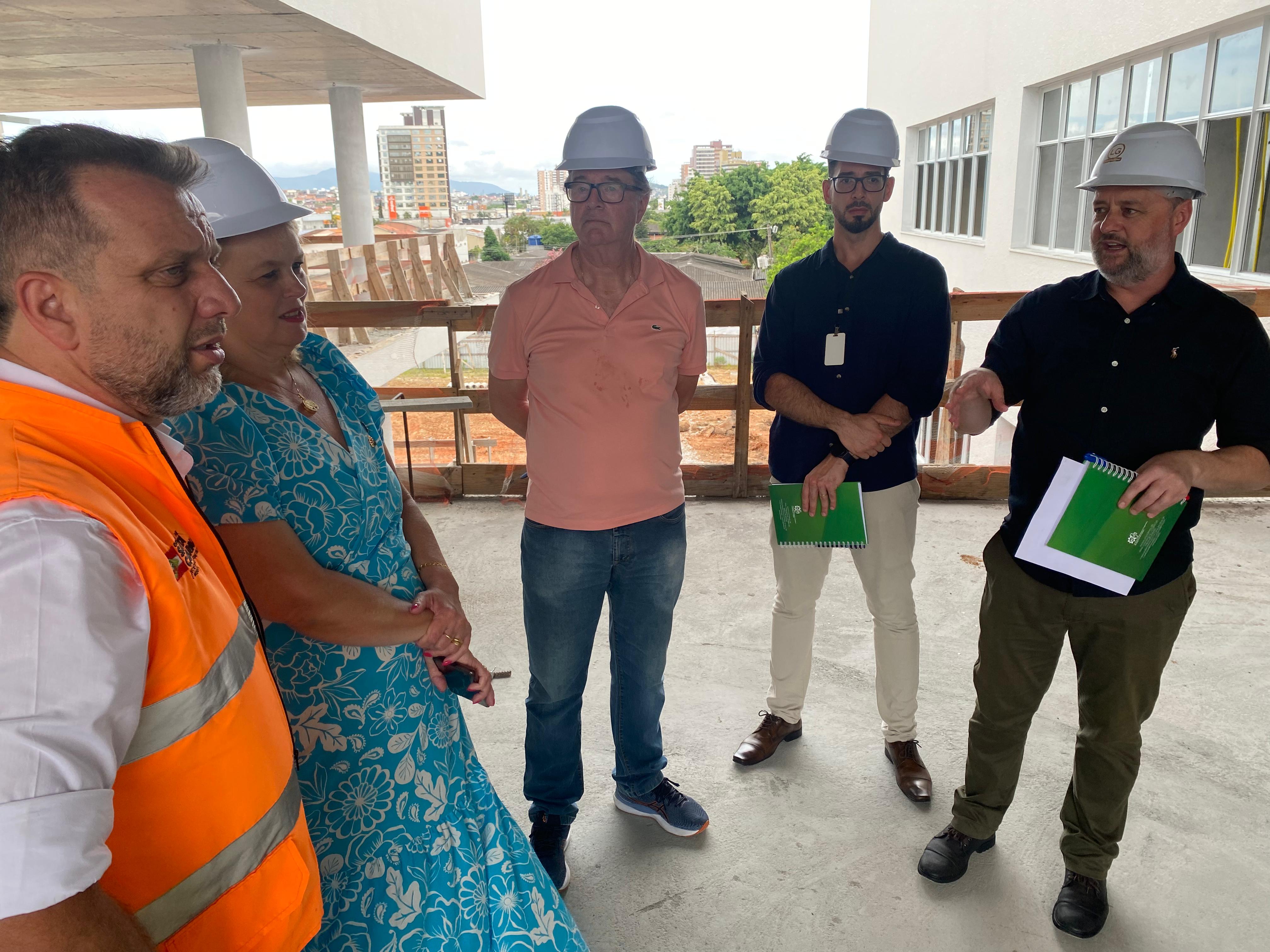 Secretário da Infraestrutura em visita à obra do novo prédio da FCEE cinco pessoas em pé, com capacetes de proteção, conversam dentro de obra