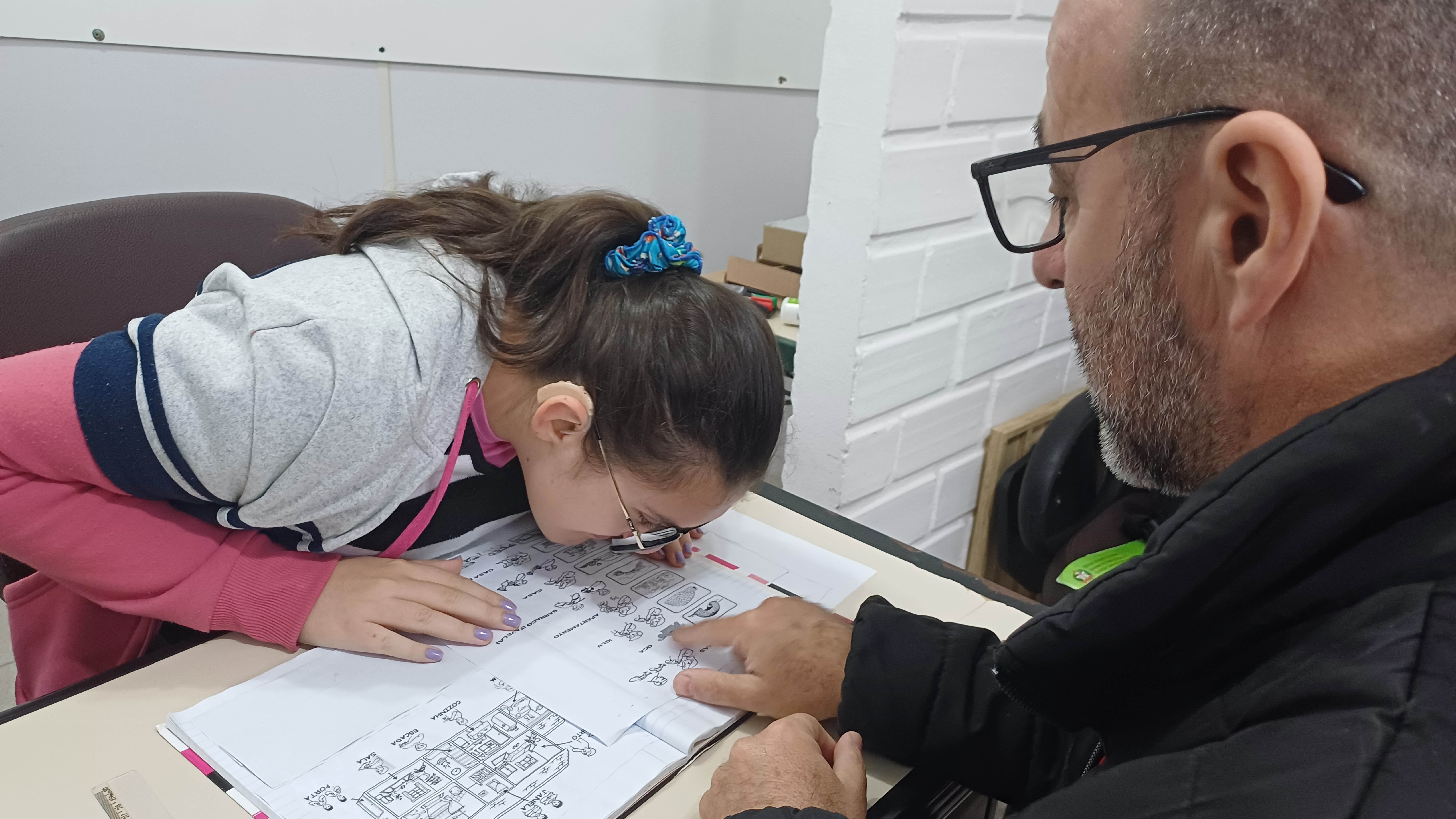 Atendimento educacional especializado na FCEE Duas pessoas sentadas em mesa de estudo, um professor e uma menina