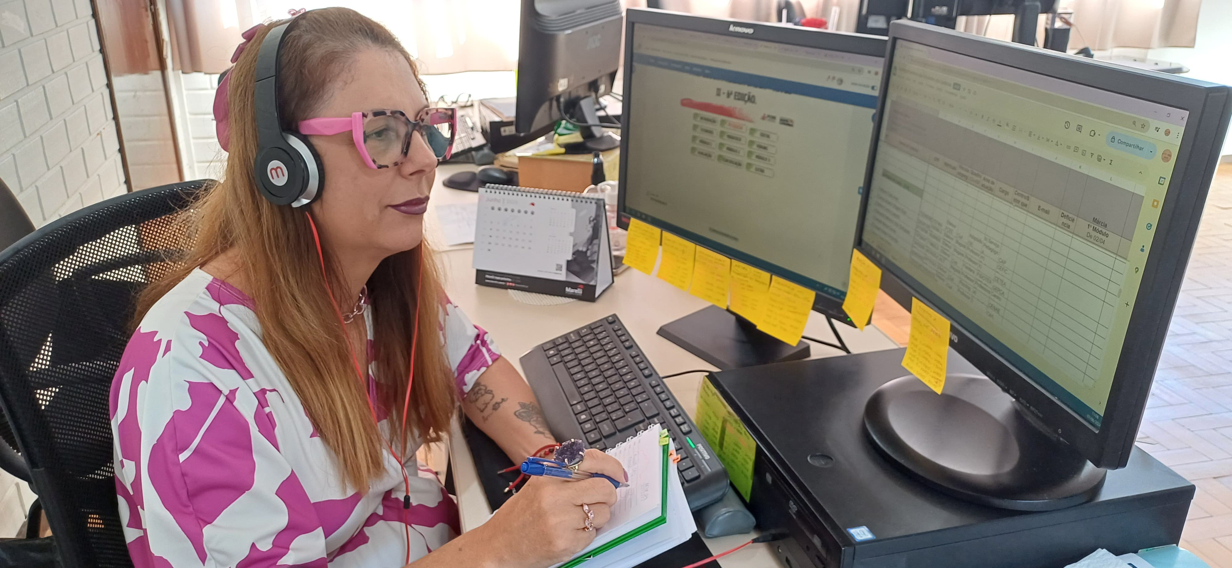 Pesquisadora Márcia Cristina Martins em ação na Gerência de Capacitação da FCEE Mulher sentada em frente a computador com duas telas, usa fones de ouvido e faz anotações.