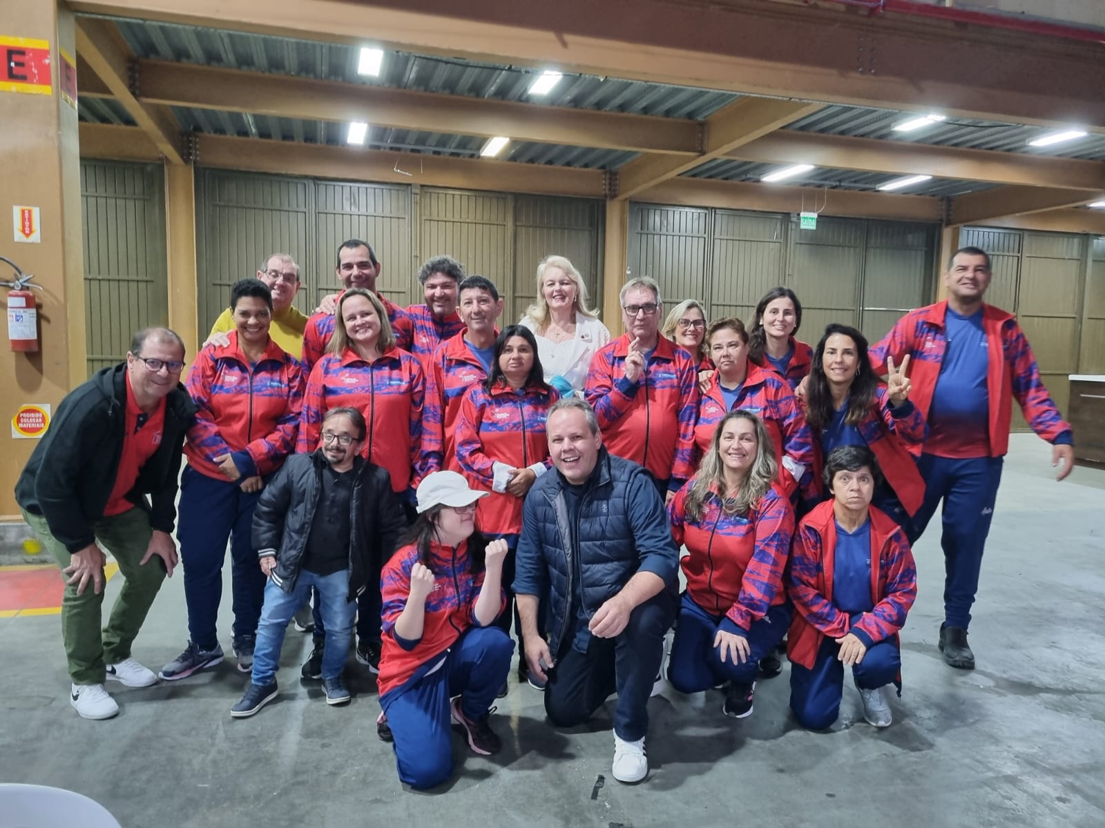 Um grupo de pessoas posam para a foto. Usam casaco esportivo vermelho. Quatro pessoas estão agachados à frente, os demais estão em pé. Estão em um galpão amplo