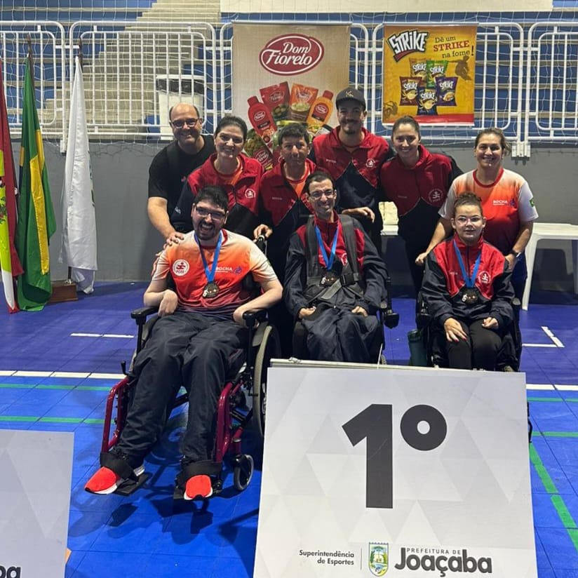 Um grupo de nove pessoas posa para uma foto no pódio de uma competição. A frente três atletas cadeirantes estão com medalhas no peito. Estão de blusa vermelha e calça preta, que fazem parte do uniforme. Atrás deles está a equipe de professores e auxiliares. A frente, em primeiro plano há número 1, em alusão ao pódio de primeiro lugar. Atrás deles, na parede do ginásio, há cartazes de várias empresas patrocinadores