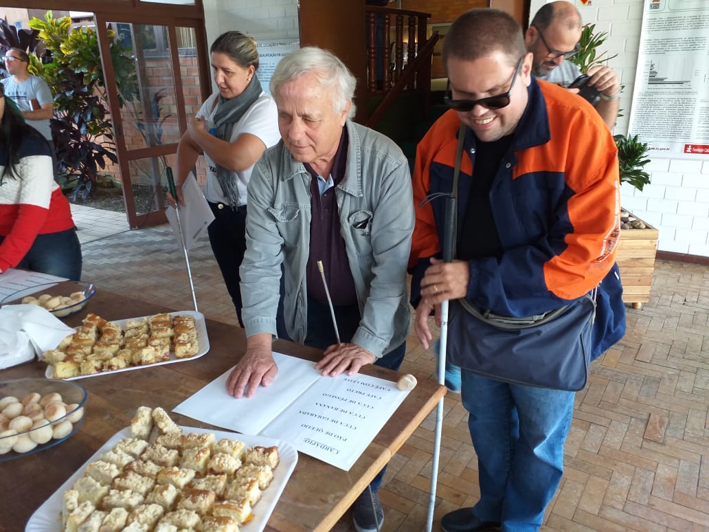 Cardápio em Braille é usado por cegos em evento na FCEE. Divulgação/FCEE. Dois homens em frente a mesa com salgadinhos, um deles usa bengala e óculos escuro. Tocam com as mãos um cartão.