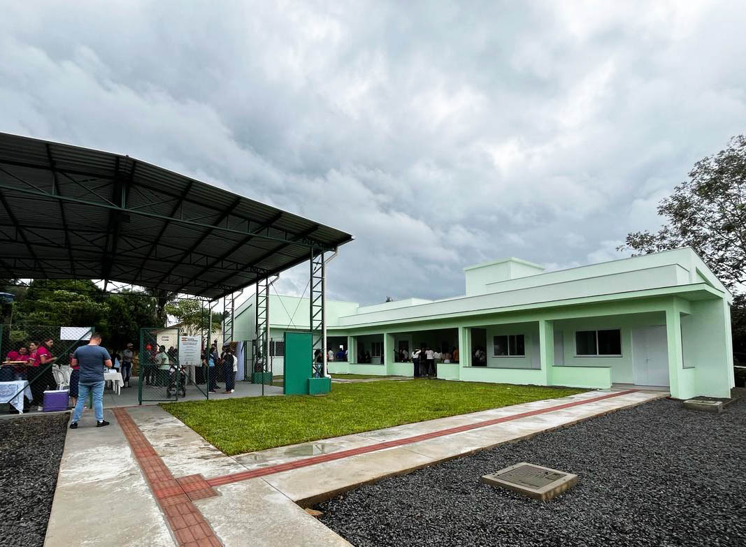 Área reformada na Apae de Água Doce Vista de prédio térreo cor verde claro ao fundo, e na frente uma quadra coberta. Piso com grama, com caminhos com piso táctil e brita, pessoas reunidas em pé dentro da quadra. Céu nublado ao fundo.
