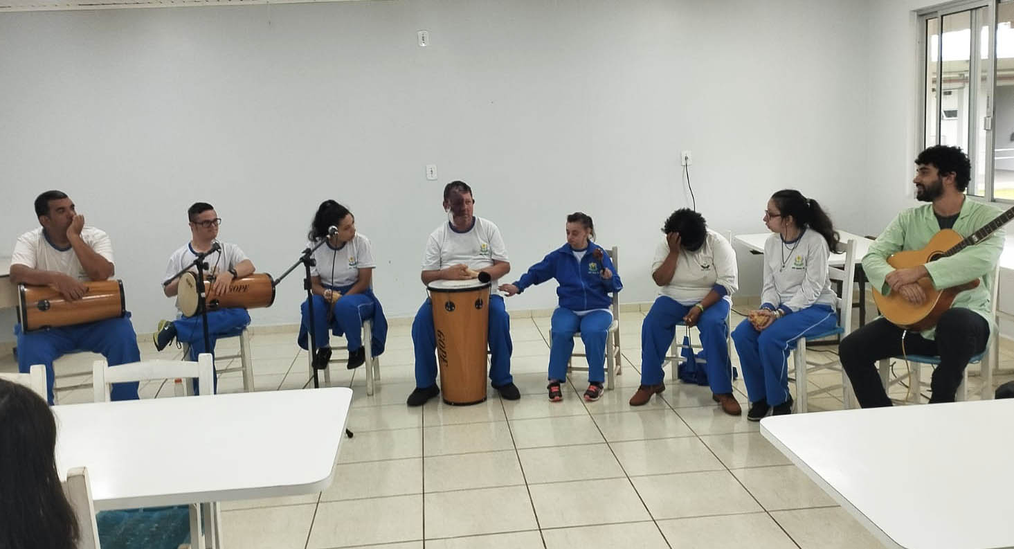 Apresentação na APAE de Joaçaba Ambiente interno, sala de aula, oito pessoas sentadas fazem apresentação de música, seguram tambores, violão e outros instrumentos, microfones com pedestal. Sete deles usma uniforme calça azul e camiseta branca.