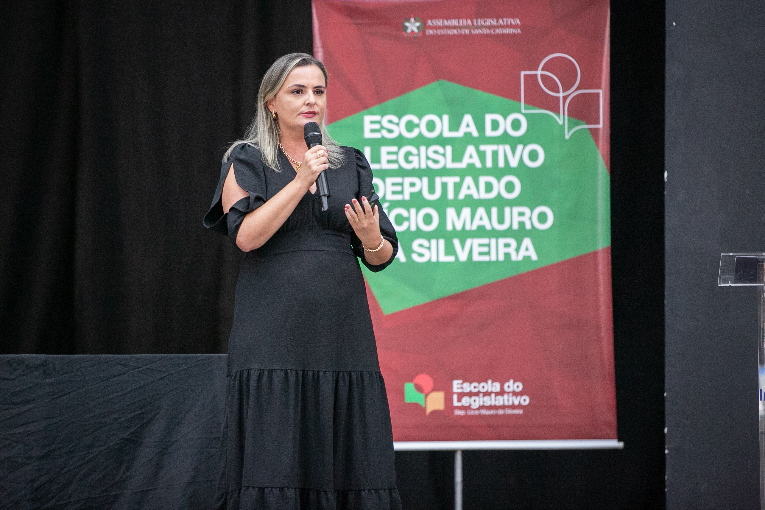 Palestrante da FCEE no seminário 1 mulher loira com vestido longo preto fala ao microfone, ao fundo banner escrito Escola do legislativo Deputado Lúcio Mauro da Silveira.