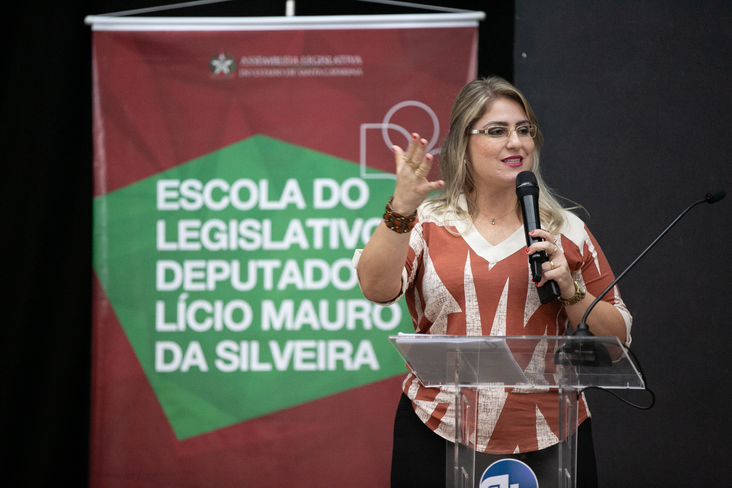 Palestrante da FCEE no seminário 1 mulher de óculos, loira cabelos lisos e pelo ombro, vestida de saia preta e blusa bege e marrom, fala ao microfone. ao fundo banner escrito Escola do legislativo Deputado Lúcio Mauro da Silveira.