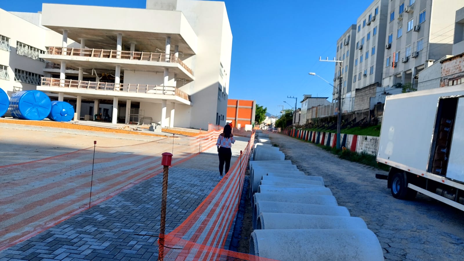 A foto mostra uma pessoa ao centro. Trata-se de uma mulher andando em um acesso a pedestres com telas protetivas. Do lado esquerdo há um prédio branco de três andares em construção. Do lado direito há tubos em concreto no meio da rua. Ao lado aparece um caminhão estacionado e prédio. A imagem registrada durante o dia e o céu está azul.