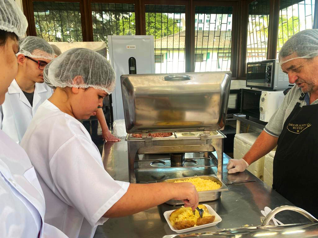 Aprendizes do CENET em aula de gastronomia cinco pessoas com toucas e avental de gastronomia, menina segura prato de comida, professor orienta, cozinha industrial.