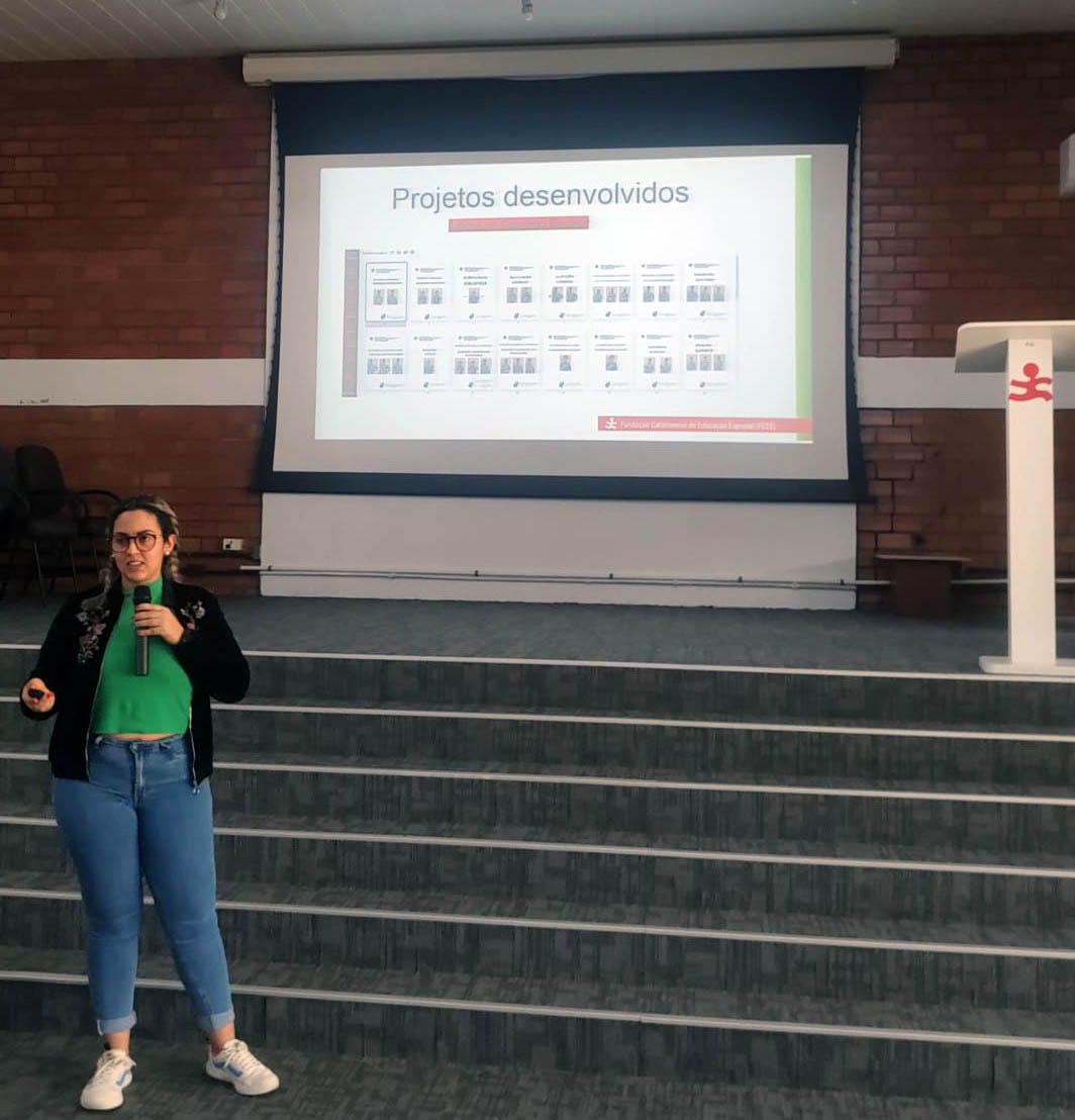 Professora Bruna Maiato Mulher fala ao microfone a frente de palco