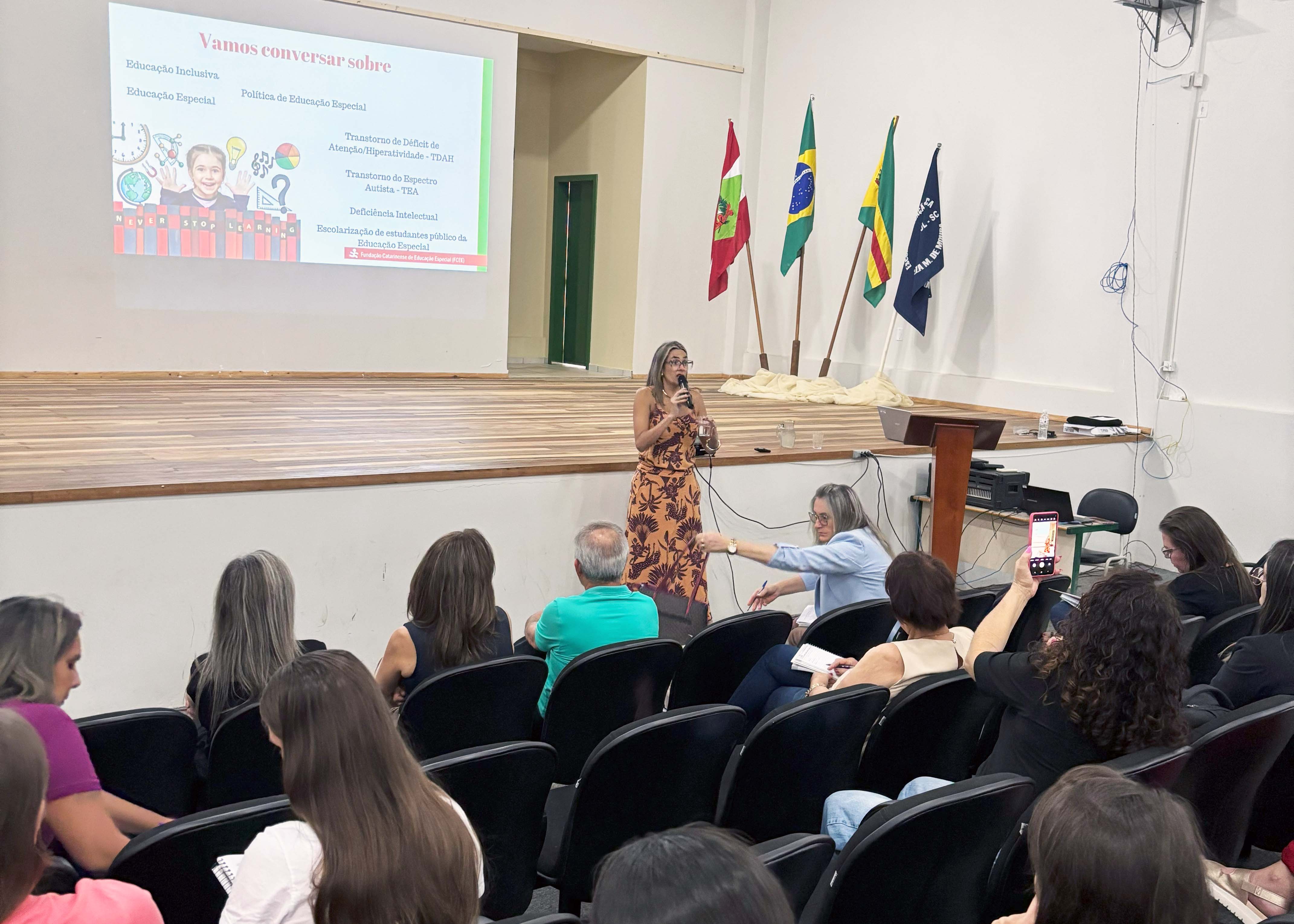auditório com pessoas na plateia, ao fundo mulher fala ao microfone