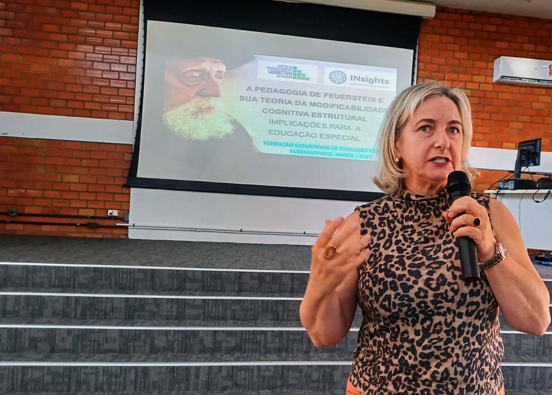 Mulher fala ao microfone em auditório, ao fundo telão